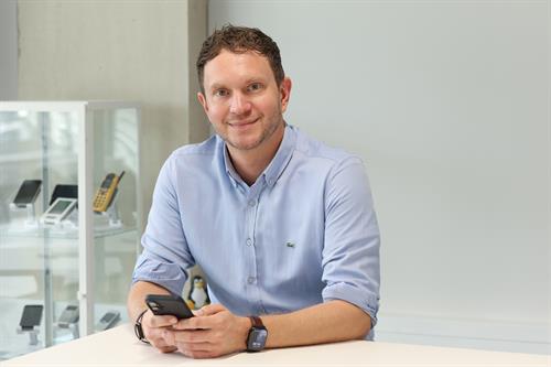 Prof. Philipp Krieter vom Fachbereich Medien der Hochschule Düsseldorf sitzt an einem Tisch und hält ein Smartphone in den Händen. Er trägt ein hellblaues Hemd und blickt lächelnd in die Kamera. Im Hintergrund ist eine Glasvitrine mit verschiedenen Mobiltelefonen zu sehen.